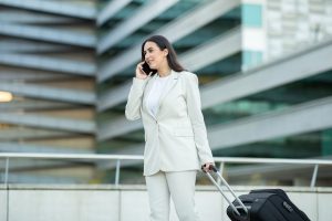 A woman in a white suit walks while talking on her cell phone, pulling a black rolling suitcase behind her