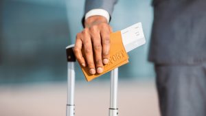 Worried woman sitting on bed, holding smartphone and passport with suitcase nearby