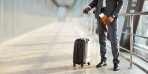 African American businessman wearing suit standing at modern airport with his suitcase, holding passport and flight tickets in his hand, cropped, panorama with copy space