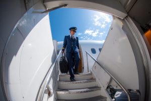 A pilot in a blue uniform walks down the steps of an airplane, holding a bag and a briefcase.