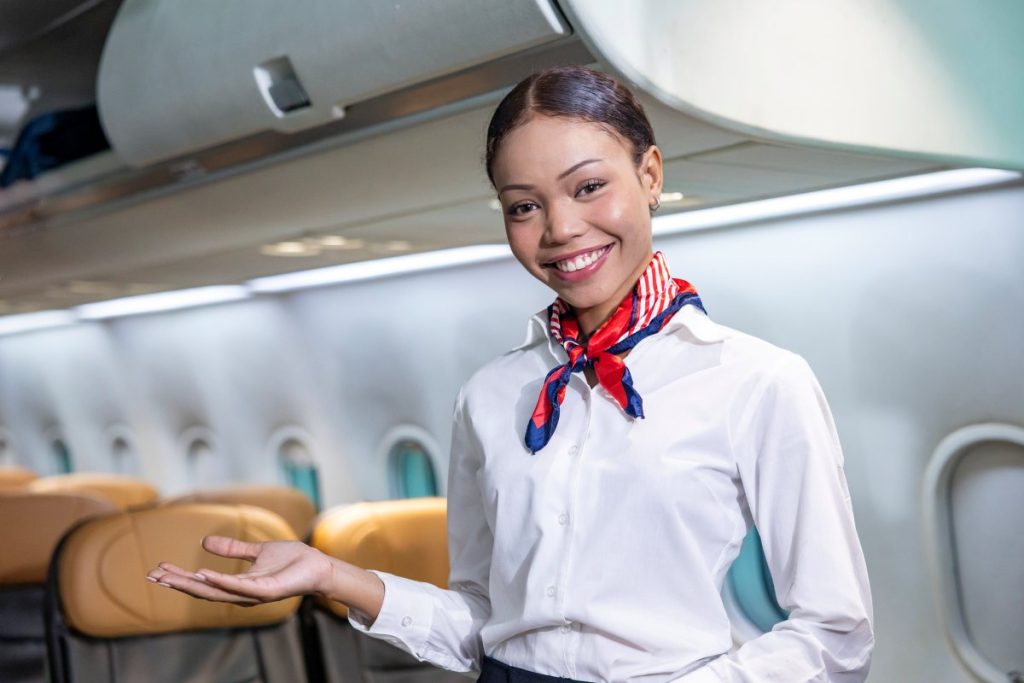 African flight attendant posing with smile face at middle of the aisle inside aircraft for welcoming passenger on board with seat on background