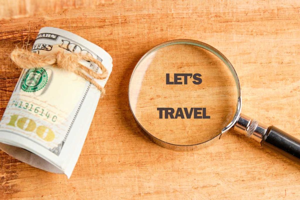 A roll of United States currency tied with twine sits next to a magnifying glass, displaying the words Lets Travel, all on a light brown wooden surface