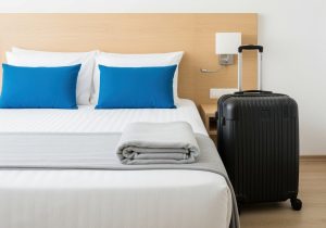 Black suitcase standing next to a pristine hotel bed with bright blue cushions